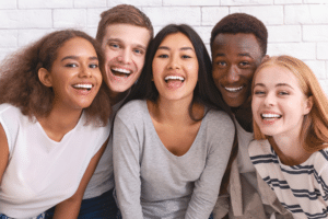 Diverse group of five smiling teenagers leaning close together in front of a white brick wall